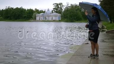 女孩站在湖边公园的雨伞下，在电话里拍照。慢动作。1920x1080。满的